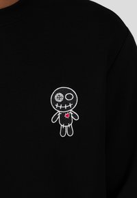 Sudadera negra con un pequeño personaje dibujado bordado con cuerpo, detalles de costura y un corazón rojo en el pecho.