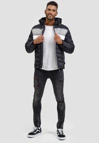 Chaqueta negra acolchada con franjas horizontales grises y blancas, cierre de cremallera y capucha. Lleva puesta una camiseta blanca y jeans negros desgastados.