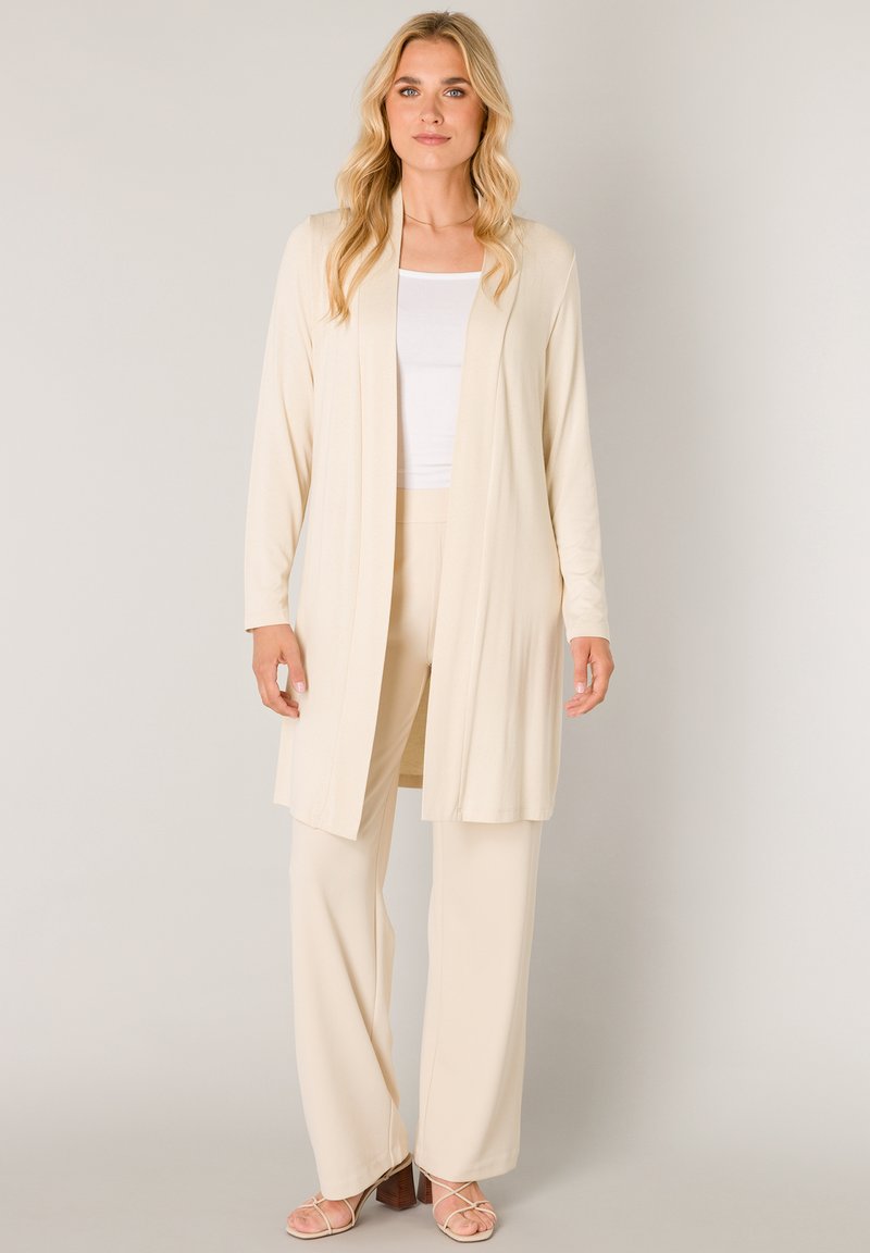 Beige cardigan met open voorkant en lange mouwen, gedragen over een wit tanktop en bijpassende beige wijde broekspijpen. Zachte stof met een gladde textuur.