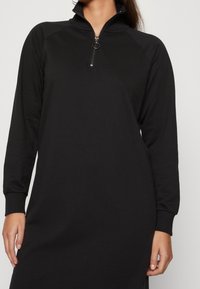 Schwarzes Langarmkleid mit hohem Kragen und Reißverschluss, aus glattem Stoff gefertigt. Mit gerippten Bündchen und gerader Silhouette.