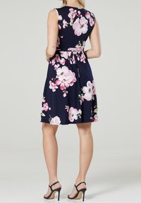 Kvinna i en ärmlös marinblå klänning med stora rosa blommor och svarta högklackade sandaler, stående med händerna på höfterna, vänd bort.