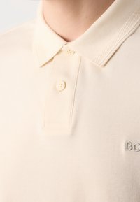 Gros plan sur un polo couleur crème avec deux boutons fermés et un logo "BOSS" brodé partiellement visible sur la poitrine.