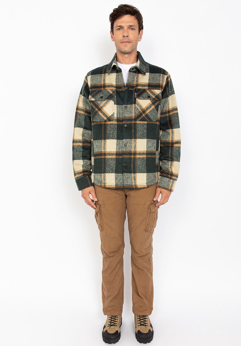 Camicia di flanella a quadri verde e beige con due tasche sul petto, abbinata a pantaloni cargo marroni e stivali impermeabili.