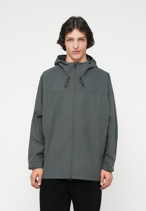 Jeune homme portant une veste zippée à capuche gris foncé avec des cordons, se tenant devant un fond blanc uni.