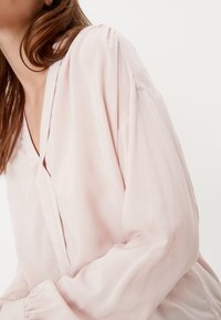 Blouse rose clair avec un col en V, dotée de manches bouffantes et amples et d'une texture lisse et légèrement brillante. Design minimaliste, sans ornements visibles.