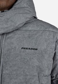 Zaklep blizu osebe, ki nosi svetlo sivo prešito jakno z visokim ovratnikom in črnim logotipom "PEGADOR" na prsih.
