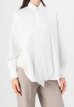 Kobieta ubrana w białą bluzkę z długim rękawem i dużą kokardą z przodu, w połączeniu z jasnobeżowymi spodniami, pokazana od szyi do bioder.