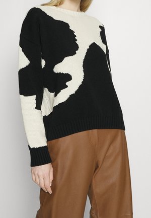 Vrouw die een zwart-wit gebreide trui met patroon en hoge bruine leren broek draagt, staand voor een effen achtergrond.
