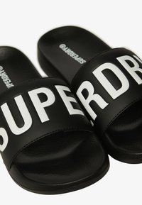 Zwarte slidesandalen met brede banden en opvallende witte "SUPERDRY" opdruk. Gemaakt van glad synthetisch materiaal met geprofileerde zolen.
