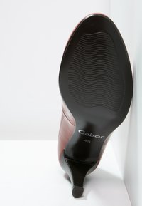 Chaussure à talon haut rouge avec une finition brillante et un bout pointu. Semelle en caoutchouc noire avec des rainures horizontales ; logo de la marque visible sur le talon.