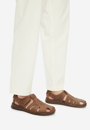 Brązowe skórzane sandały z zakrytymi palcami i zapięciem na rzepy, noszone z białymi spodniami o prostych nogawkach na jasnym tle.