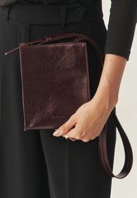 Bordo torbica za nošenje preko tijela od kože s glatkom teksturom, pravokutnog oblika, podesivim remenom i diskretnim logotipom sprijeda.