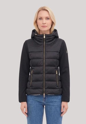 Donna con capelli biondi che indossa una giacca trapuntata nera con cappuccio e dettagli di zip dorati, abbinata a jeans blu, in piedi davanti a uno sfondo semplice.