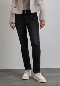 Osoba v černých skinny džín ách, béžovém kratším kabátu a bílých teniskách, stojící na šedé podlaze proti jednoduché zdi.