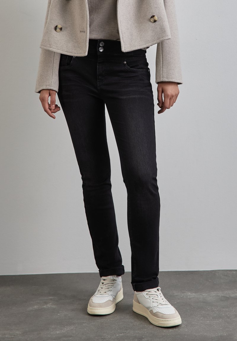 Osoba v černých skinny džínách, béžovém kratším kabátu a bílých teniskách, stojící na šedé podlaze proti jednoduché zdi.