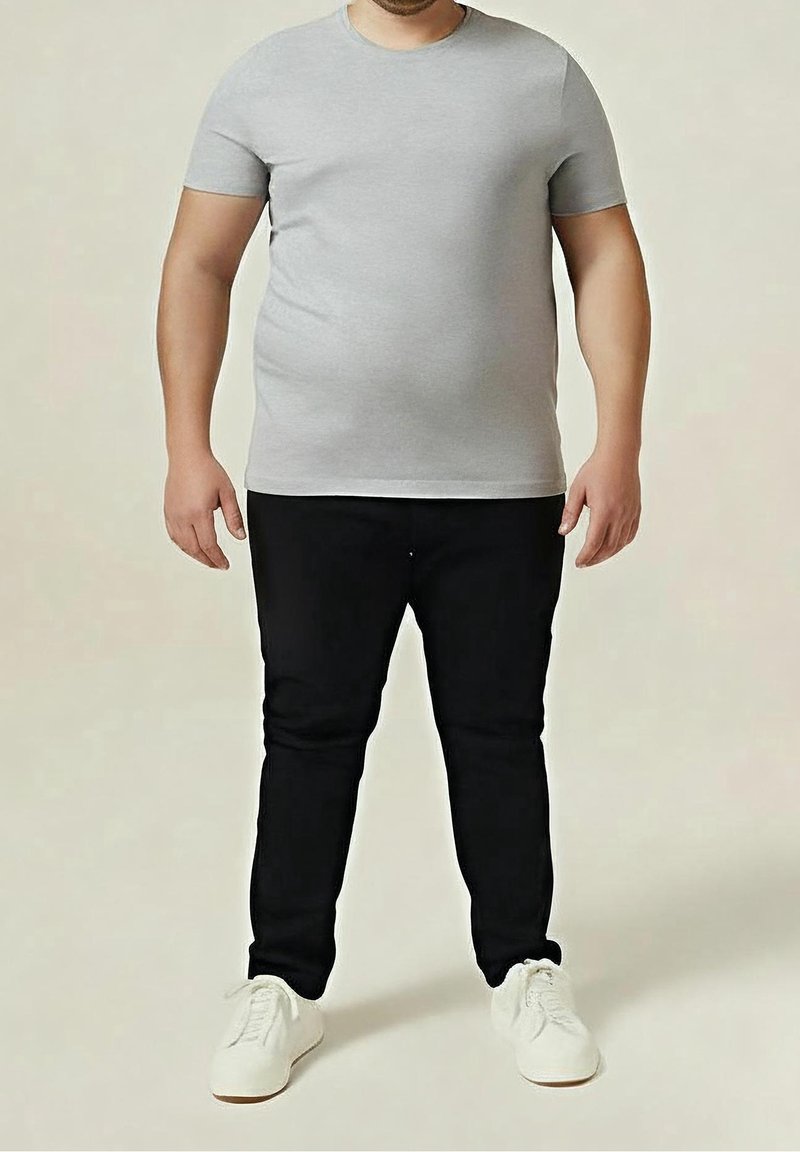 Homme debout portant un t-shirt gris à manches courtes, un pantalon noir et des baskets blanches sur un fond neutre uni.