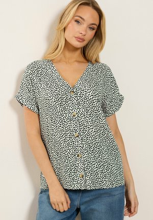 Frau trägt eine kurzärmelige weiße Bluse mit schwarzen Punkten und Knöpfen vorne, kombiniert mit blauen Jeans.