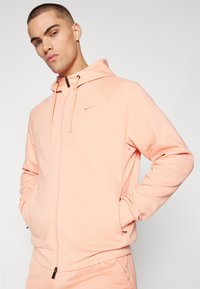 Lys rosa zip-up hettegenser med snorhette, svart glidelås og en liten gulllogo på brystet. Glatt, myk stoff med ribbede mansjetter.