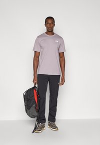 Ανοιχτό μωβ κοντομάνικη μπλούζα με λευκό λογότυπο, μαύρο παντελόνι και μαύρο σακίδιο με κόκκινη εσωτερική επένδυση. Τα παπούτσια έχουν μαύρο και μπεζ σχέδιο.