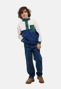 Pull en polaire bicolore en blanc, vert et bleu, avec un col haut, une fermeture éclair et une poche avant ; associé à un jean en denim ample et des chaussures marron.