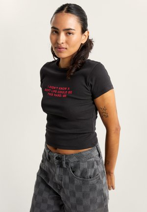Zwart aansluitend T-shirt met rode tekst in het midden. Gedragen met hoog getailleerde grijze geruite broek. Soepele stof, korte mouwen en ronde hals.