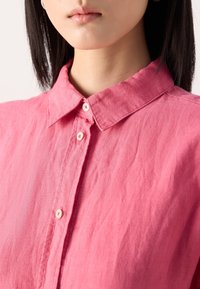 Femme portant une chemise en lin rose vif à boutons avec col, montrée du cou aux épaules.