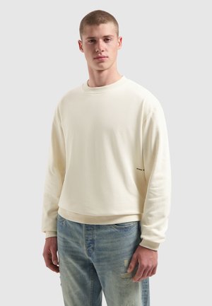 Jonge man in een crèmekleurig sweatshirt met ronde hals en lichtblauwe versleten spijkerbroek, staand tegen een effen lichtgrijze achtergrond.