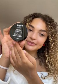 Svart cirkulär behållare med vit text märkt "NYX Mineral Matte Finishing Powder." Modellen håller produkten med händer som visar en ljus hudton.