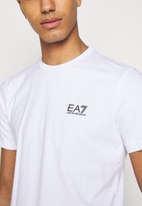 Camiseta blanca de algodón con cuello redondo, que presenta un pequeño logo negro de EA7 Emporio Armani en el área del pecho izquierdo. Textura suave.