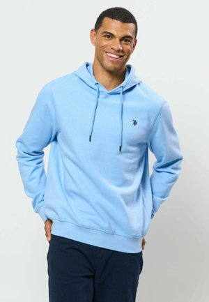 Homme souriant portant un sweat à capuche bleu clair avec cordons et un pantalon foncé, debout devant un fond clair uni.