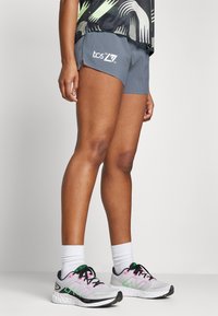 Dame grå treningsshorts med perforert design og hvitt grafisk logo, kombinert med lette løpesko i grått, rosa og grønne detaljer.