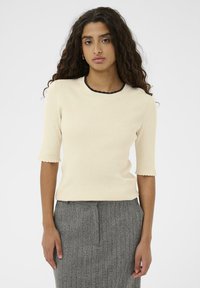 Ung kvinde med langt curly hår iført en cremefarvet ribbet kortærmet top og en grå struktureret nederdel, der står op ad en ensfarvet hvid baggrund.