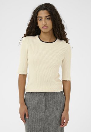 Ung kvinde med langt curly hår iført en cremefarvet ribbet kortærmet top og en grå struktureret nederdel, der står op ad en ensfarvet hvid baggrund.