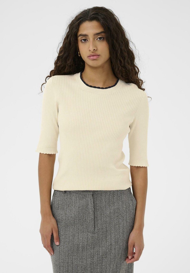 Ung kvinde med langt curly hår iført en cremefarvet ribbet kortærmet top og en grå struktureret nederdel, der står op ad en ensfarvet hvid baggrund.