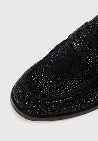 Svart slip-on sko i texturerat material, prydd med små, glänsande kristaller. Har en rundad tå och en slät kant runt sulan.