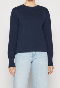 Felpa a maniche lunghe navy con design floreale ricamato lungo il collo. Tessuto morbido, vestibilità rilassata e polsini a costine. Indossata con jeans azzurri.