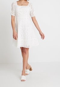 Robe blanche avec un décolleté carré et des manches bouffantes, présentant des motifs découpés et une jupe évasée. Portée avec des mules blanches à talons.