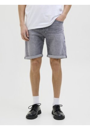 Homme portant un short en denim gris retroussé, des chaussettes blanches, des chaussures noires et un T-shirt blanc, une main dans la poche.