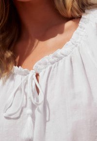 White blouse with a ruffled neckline, featuring a tie closure with a tassel accent. The fabric appears lightweight and textured.