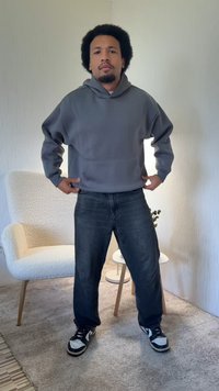 Szara bluza z kapturem wykonana z miękkiego materiału, o luźnym kroju; w połączeniu z ciemnymi dżinsami i czarno-białymi trampkami. Prosty wzór bez widocznych wzorów.