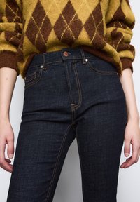 Ciemnoniebieskie dżinsy z pomarańczowymi przeszyciami, wysoki stan, pięć kieszeni. Noszone z żółtym swetrem w brązowy wzór.