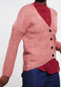 Textureret pink cardigan med lange ærmer, V-hals og fem sorte knapper. Båret over en rød skjorte og stylet med blå bukser.