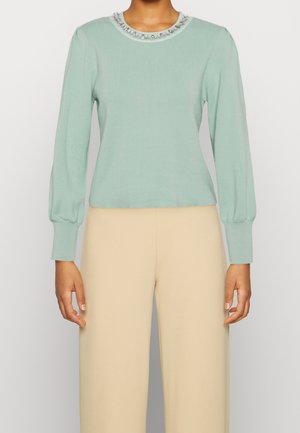 Femme portant un pull à manches longues vert pastel avec des ornements en perles et perles au niveau de l'encolure et un pantalon beige taille haute.