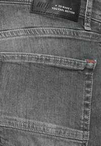 Šedé džínsy z denimu se zadní kapsou, s texturovaným povrchem, prošitými švy a černou koženou etiketou s bílým textem. Viditelný červený akcent.