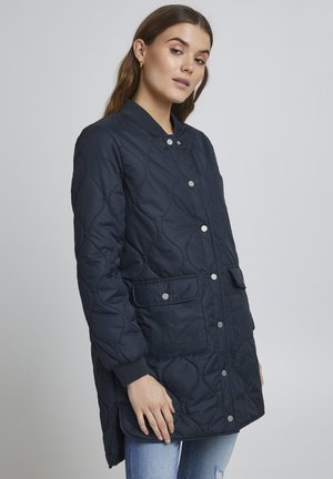 Marineblau gesteppte Jacke mit Druckknopfverschluss, Rippbündchen, zwei vorderen Taschen und abgerundetem Saum. Das Material wirkt glatt und hat eine weiche Textur.
