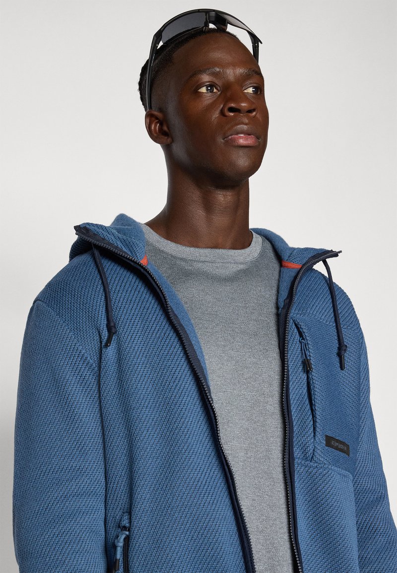 Blau strukturierte Zip-Hoodie mit Kapuze, grauem Rundhals-T-Shirt darunter und einer kleinen Brusttasche. Sonnenbrille liegt auf der Kapuze.