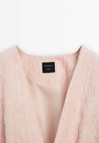 Veste en tissu rose clair texturé avec une doublure intérieure lisse et une étiquette noire portant l’inscription Massimo Dutti Studio à l’intérieur du col.