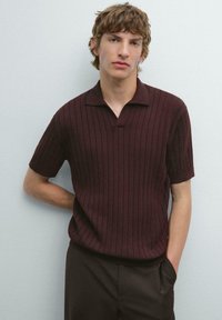 Kortärmad polo t-shirt i burgunderröd med vertikal ribbad textur, krage och avslappnad passform. Kombinerad med mörka byxor.