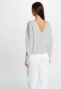 Maglione grigio in maglia con scollatura a V profonda, caratterizzato da dettagli con bottoni lungo la spina dorsale e maniche lunghe. Abbinato a pantaloni bianchi a gamba larga.
