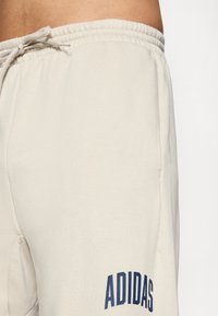 Imagem em close-up de uma pessoa a usar calças de treino beje claro da Adidas, com o logotipo azul-marinho na coxa e o cordão da cintura amarrado em forma de laço.